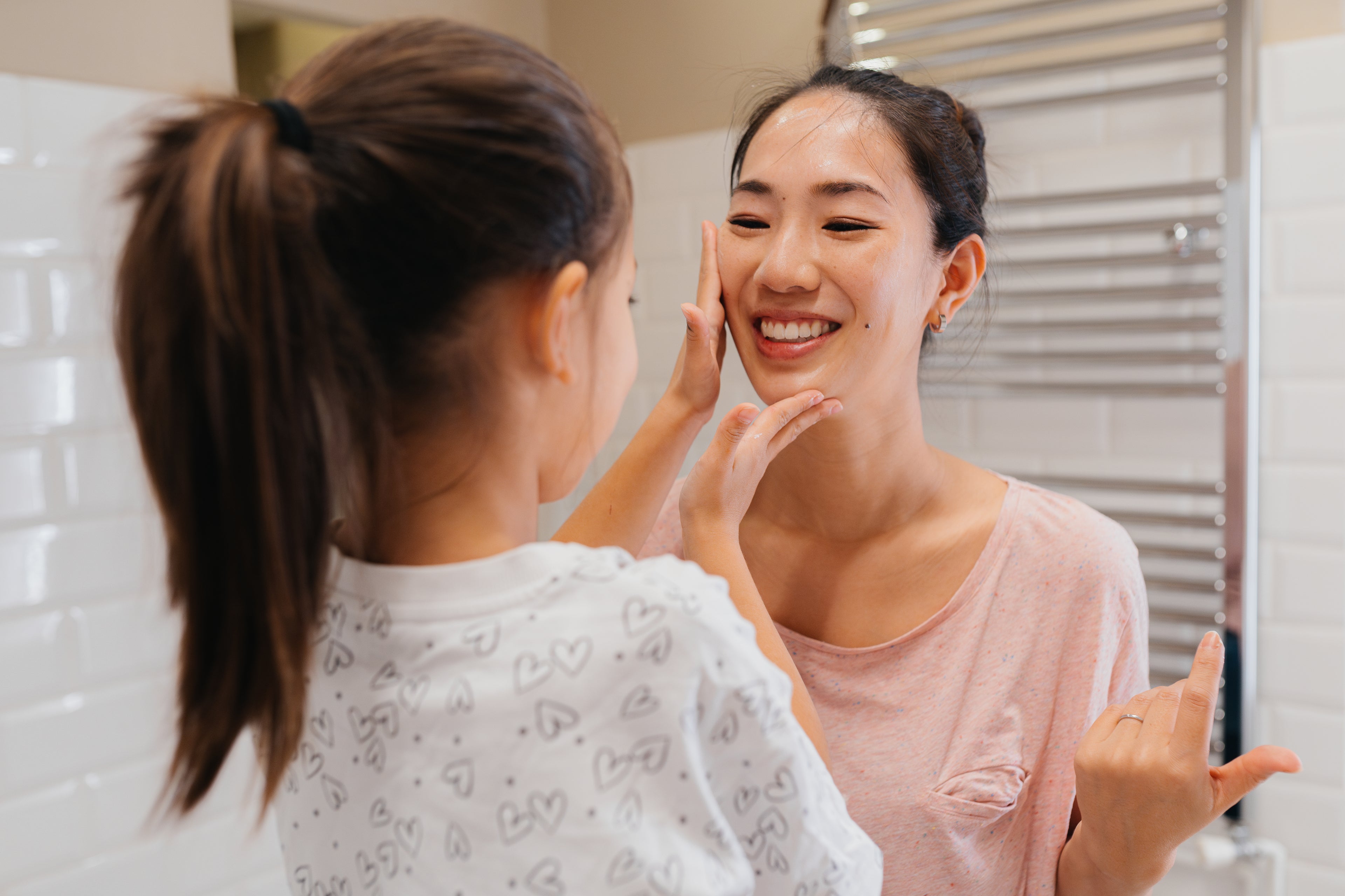 A mother is smiling at her daughter who is applying a skincare cream to her mother's face. They are standing in a bright, white bathroom.