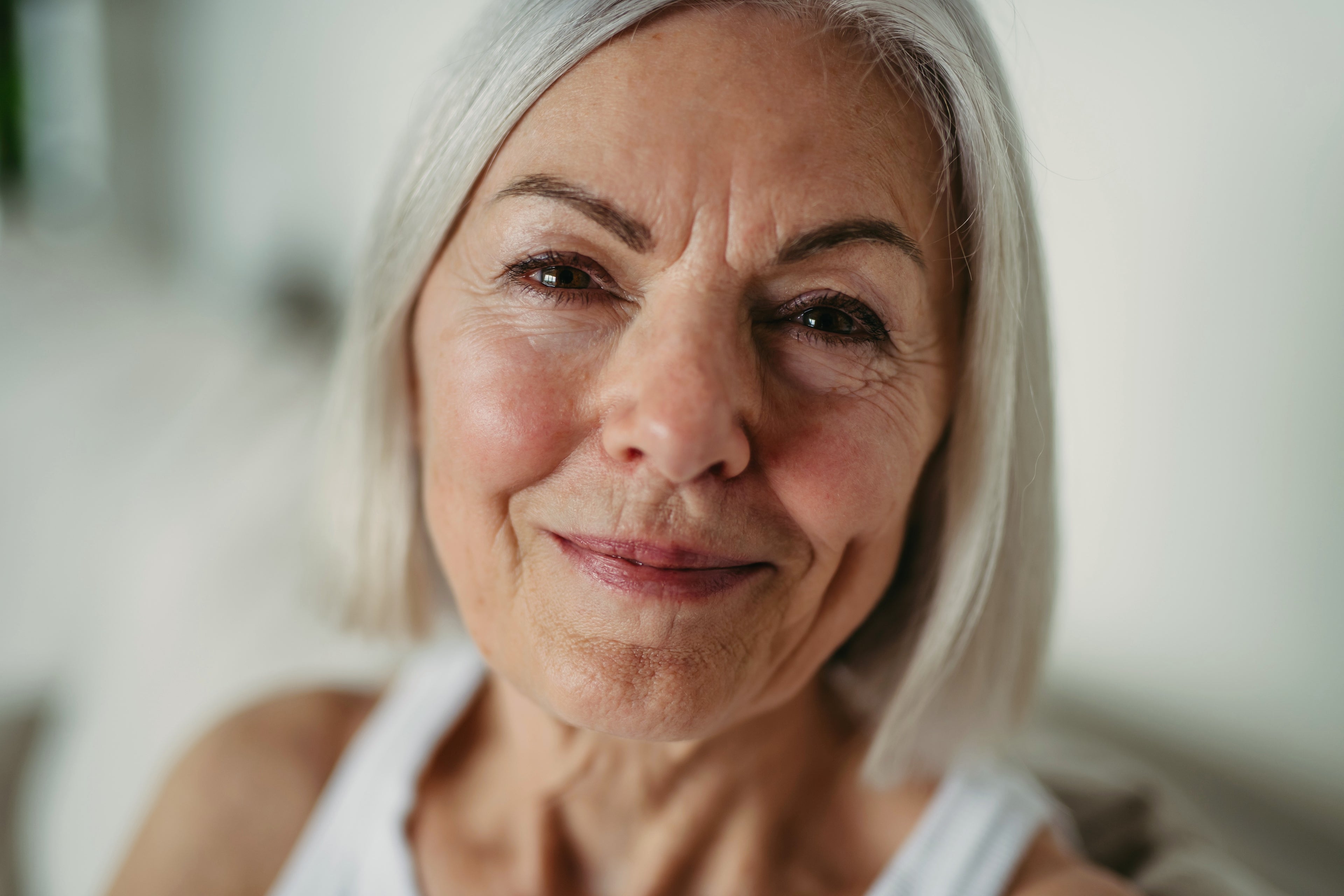 A woman in her 50s with natural, beautiful, aging skin and a short grey bob is looking into the camera smiling. Her skin is happy and healthy.